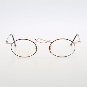May include: Oval spectacles with a gold-coloured bridge and black temples. The rims have a colourful pattern of red, green, and yellow. The text "TAZI MADE IN JAPAN" is visible on the left lens.