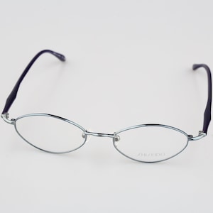 May include: Oval-shaped eyeglasses featuring a thin, metallic silver frame and dark purple temple arms. The right lens displays the brand name SHISEIDO printed in a subtle, light-colored font. These glasses have a bridge and clear lenses.