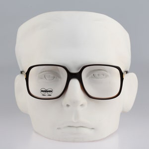 May include: Black framed glasses with brown tinted lenses. The glasses have a gold accent on the frame and the word "MORWEH" printed on the right lens.