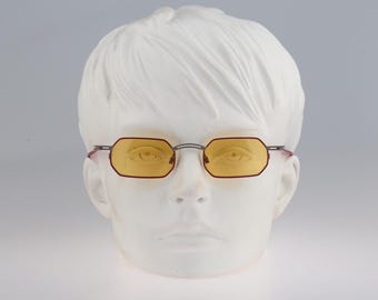 May include: Eyeglasses featuring thin, dark metal frames with a distinct octagonal shape and translucent yellow-tinted lenses. The glasses are displayed on a white mannequin head, showcasing the slim bridge and slender temples of the eyewear.