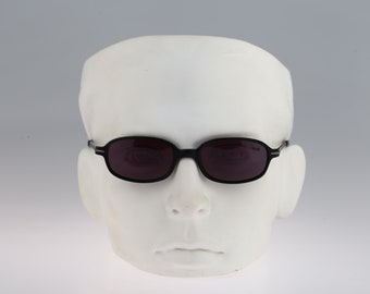 May include: Black oval-shaped sunglasses with dark lenses on a white mannequin head.