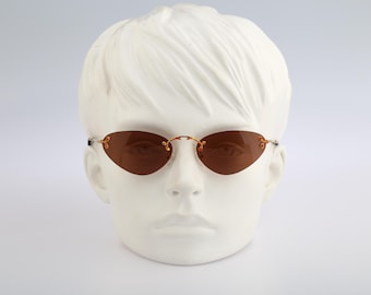May include: Rimless sunglasses featuring oval-shaped brown tinted lenses and thin gold-tone metal temples. The bridge and hinges display a delicate filigree-inspired metal design, connecting the lenses without a full frame structure.