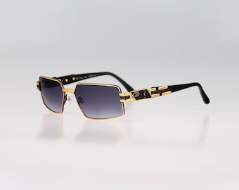 Puede incluir: Gafas de sol rectangulares con montura de metal dorado y detalles en negro. Las lentes tienen un tono degradado gris oscuro. Las patillas presentan un diseño decorativo en dorado y negro con un pequeño emblema de logo cerca de las bisagras.