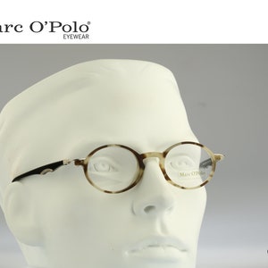 Può includere: Occhiali rotondi con montatura a fantasia tartarugata, aste nere e lenti trasparenti. Gli occhiali sono esposti su una testa di manichino bianca. Il marchio "Marc O'Polo" è visibile in alto.