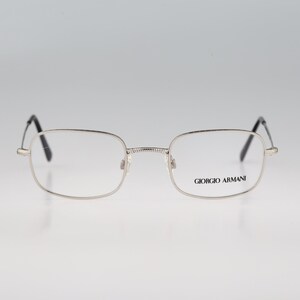 May include: Silver-toned metal spectacles with rectangular frames and thin temples. The right lens features the text GIORGIO ARMANI printed in black. The bridge has a textured, ribbed design, and the temple tips are finished in black.