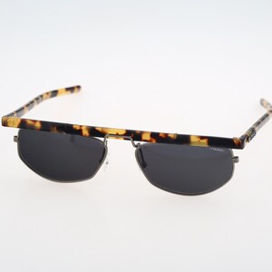 Könnte beinhalten: Sonnenbrille mit flacher Oberkante und einem braun-schwarzen Schildpattmuster. Die Gläser sind dunkelgrau und oval geformt, eingefasst in einen dünnen Metallrahmen. Die Bügel weisen ebenfalls das Schildpattmuster auf und bilden eine Einheit.
