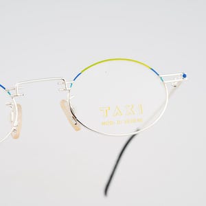 May include: Silver-framed eyeglasses with round lenses. The top of the frames are accented with a thin line of green and blue paint. The text "TAXI MODI DI VEDERE" is printed on the right lens.