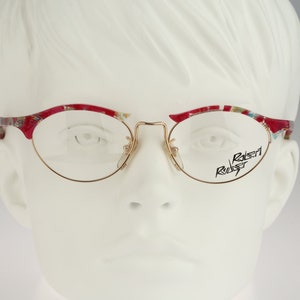 Puede incluir: Gafas ovaladas de color rojo y dorado con un patrón floral en el marco. El nombre de la marca "Robert Robert" está impreso en la lente derecha.