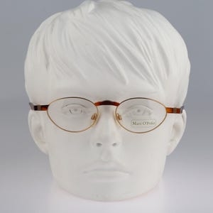 May include: Oval-shaped spectacles with a thin, metallic gold frame and brown tortoiseshell-patterned accents on the bridge and temples. The right lens features the brand name Marc O'Polo printed in black text on the clear glass surface.