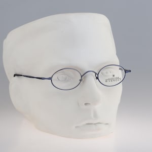 Puede incluir: Gafas de forma ovalada con una montura fina de metal oscuro. El cristal derecho presenta el texto LUNETTES GIVENCHY impreso en negro. Las gafas se muestran sobre una cabeza de maniquí blanca, destacando las varillas y el puente finos.