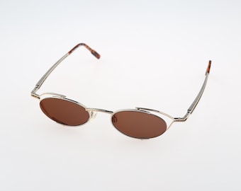May include: Oval sunglasses featuring a thin silver-tone metal frame and brown tinted lenses. The temples are slender with brown tortoiseshell-patterned tips, and the bridge connects the two oval-shaped lenses in a streamlined, minimalist design.