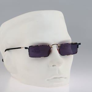 Puede incluir: Un par de gafas de sol rectangulares negras y doradas con lentes oscuros. Las gafas están en una cabeza de maniquí blanca.