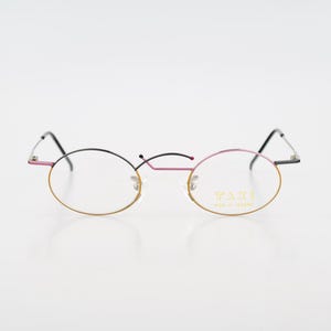 May include: Oval-shaped eyeglasses with a gold-colored frame around the lenses, transitioning to black arms. The top of the frame is pink and black. The lenses are clear, and the glasses have a delicate, minimalist design.