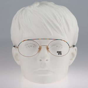 May include: Gold-rimmed round eyeglasses with a multi-colored stripe on the top of the frame. The brand name "DOLCE VITA" is printed on the right lens.