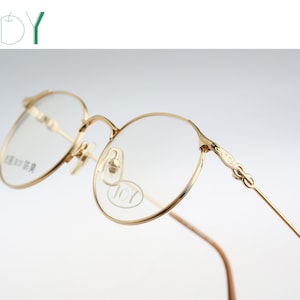 May include: Gold-tone metal eyeglasses with round frames and thin temples. The frames feature intricate engraved detailing on the hinges. The clear lenses have a small oval logo printed on the right side with the text JOY and Japanese characters.