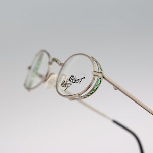 Puede incluir: Gafas redondas plateadas con detalles verdes en el marco. El nombre de la marca "Robert Rose" está impreso en la lente.