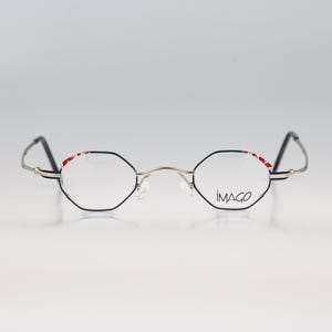 May include: Silver-toned metal spectacles with a distinct hexagonal frame shape. The upper rims feature a decorative red and white pattern. The right lens is etched with the brand name IMAGO in black lettering. The thin temples are black.