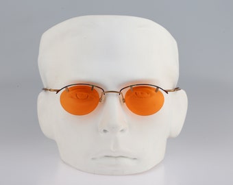 Puede incluir: Un par de gafas de sol sin montura con lentes naranjas en una cabeza de maniquí blanca. Las gafas tienen un fino marco de metal dorado.