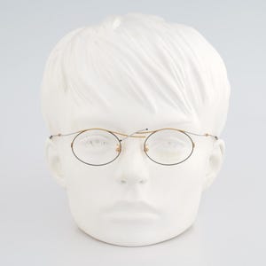 May include: Oval-shaped eyeglasses with a gold-colored frame and thin, dark lenses. The glasses are displayed on a white mannequin head. The overall style is vintage and classic.