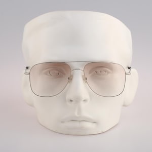 Puede incluir: Gafas de aviador con montura plateada y lentes tintadas, expuestas sobre una cabeza de yeso blanco. Las gafas tienen un doble puente y una forma rectangular clásica. Las lentes tienen un ligero tono rosado.