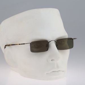 Könnte beinhalten: Rechteckige Sonnenbrille mit dunkelgrauem Metallrahmen und getönten Gläsern, präsentiert auf einem weißen Schaufensterkopf. Die Bügel haben ein Schildpattmuster. Die Sonnenbrille hat einen klassischen Stil.