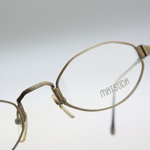 Può includere: Occhiali in metallo color oro antico con montatura di forma ovale e dettagli incisi complessi sul ponte e sulle aste. La lente destra presenta il nome del marchio MATSUDA stampato in un carattere nero sans-serif sul vetro trasparente.