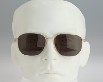May include: Sunglasses with a thin gold-tone metal frame and dark brown tinted lenses. The frames feature a rounded rectangular shape with a slim bridge and thin temples, providing a classic look. The lenses are dark and opaque, offering full coverage.