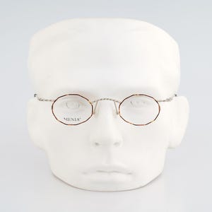 May include: Oval-shaped eyeglasses with a tortoiseshell pattern rim and thin, twisted silver-tone metal temples. The lenses are clear, and the word MENIA is printed in black on the left lens. The frames feature a bridge with a twisted metal design.