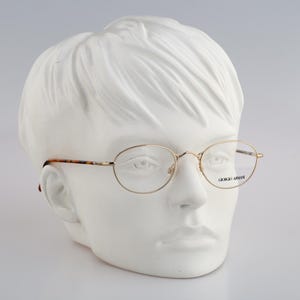 May include: Gold-rimmed eyeglasses with clear lenses on a white mannequin head. The glasses have a subtle tortoise shell pattern on the arms and the text "Giorgio Armani" is printed on the right lens.