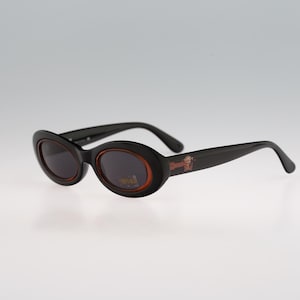 May include: Black oval sunglasses with brown frames and lenses. The sunglasses have a small logo on the right frame.