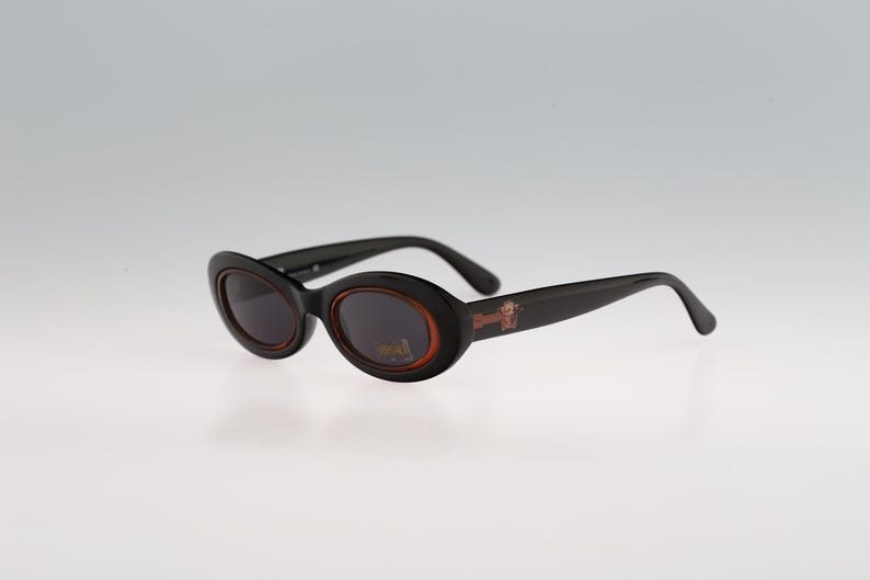 May include: Black oval sunglasses with brown frames and lenses. The sunglasses have a small logo on the right frame.