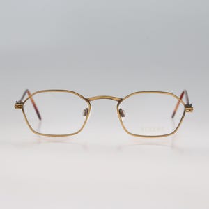 May include: A pair of spectacles with a thin, metallic gold-toned frame featuring a distinct hexagonal shape. The clear lenses are held by a slender bridge and temples that transition into dark, tortoiseshell-patterned tips at the ends.
