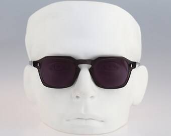 May include: Dark charcoal gray sunglasses with a distinct geometric, angular frame shape and dark tinted lenses. The glasses feature a keyhole bridge design and small silver-toned metal rivets on the outer corners of the front frame.