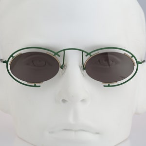 May include: Green oval-shaped sunglasses with a gold metal frame and brown lenses. The sunglasses are on a white mannequin head.