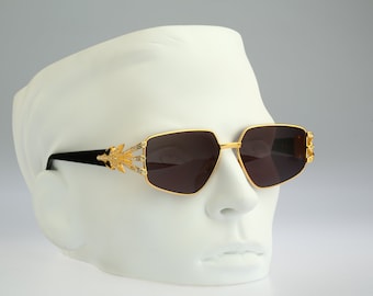 Könnte beinhalten: Goldfarbene Sonnenbrille mit schwarzen Bügeln und klaren Strasssteinen am Rahmen. Die Sonnenbrille hat eine quadratische Form und dunkle Gläser.