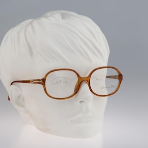 May include: Brown tortoiseshell patterned eyeglasses with a rounded rectangular frame shape. The temples feature a gold-toned metal detail with the CD logo. The right lens is etched with the text Christian Dior in a cursive script font.