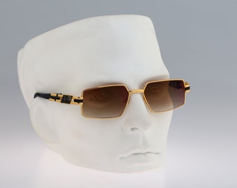 Könnte beinhalten: Goldfarbene Sonnenbrille mit schwarzen Akzenten und braunen Verlaufsgläsern. Die Sonnenbrille hat einen quadratischen Rahmen und befindet sich auf einem weißen Mannequin-Kopf.