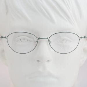 Könnte beinhalten: Oval geformte Brille mit einem dünnen, dunkelmetallischen Rahmen. Die Brille verfügt über einen Steg und Bügel, die durch kleine, sichtbare Scharniere verbunden sind. Auf dem rechten Glas befindet sich der Schriftzug prodesign in hellgrau.
