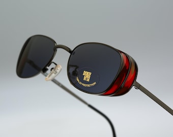 May include: Dark-tinted sunglasses with thin metal frames and side shields featuring a reddish-brown tortoiseshell pattern. A circular sticker on the right lens displays the text DOLCE VITA and 100% UV PROTECTION in gold lettering.
