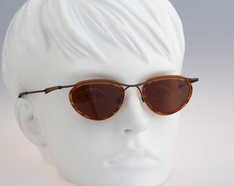 May include: Oval sunglasses featuring brown tortoiseshell rims and a thin, dark metal frame. The lenses are tinted a solid brown colour. The glasses are displayed on a white mannequin head, showcasing the slim metal temples and bridge design.