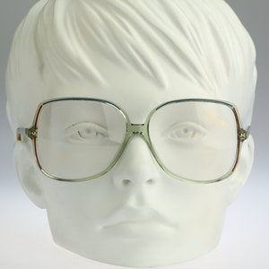 Könnte beinhalten: Eine Brille im Vintage-Stil mit einem hellgrünen Gestell und klaren Gläsern. Das Gestell hat einen dünnen, silberfarbenen Metallstreifen am Rand der Gläser.