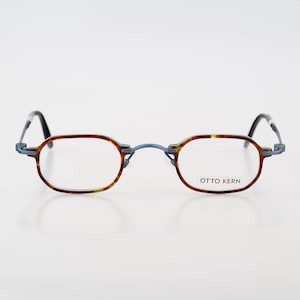 May include: Oval eyeglasses with a tortoiseshell pattern frame and a blue metal bridge. The right lens features the text OTTO KERN in black and red lettering. The temples are thin and dark, connecting to the front frame with a metal hinge.