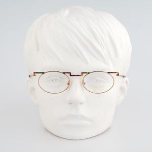 May include: Oval-shaped eyeglasses with a gold-colored frame and a colorful bridge. The glasses are displayed on a white mannequin head. The frame has a unique design with a colorful pattern on the bridge and temples.
