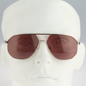 May include: Aviator-style sunglasses featuring a thin gold metal frame and a double bridge design. The lenses are a deep reddish-brown tint with a subtle tortoiseshell pattern along the top rim of the frame, providing a distinct look.