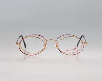 May include: Oval-shaped eyeglasses featuring a thin gold-tone metal frame with a multi-colored speckled pattern along the outer rim. The right lens displays the red text Casanova with a small circular logo. The temples are thin and dark-colored.