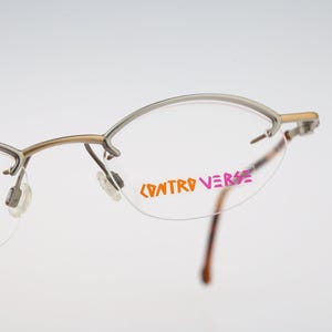 Puede incluir: Gafas semi-al aire con montura de metal dorado y lentes transparentes. El cristal derecho presenta el texto CONTROVERSE impreso en letras naranjas y rosas. El diseño incluye almohadillas nasales ajustables y varillas de metal finas.
