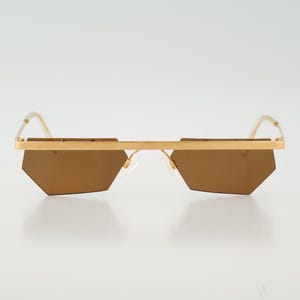 May include: Gold-framed sunglasses with geometric, brown-tinted lenses. The sunglasses have a straight brow bar and thin temples. The lenses are a unique, angular shape, and the overall design is modern and stylish.