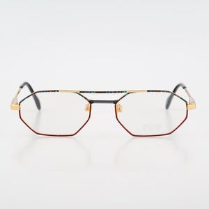Puede incluir: Gafas geométricas con puente dorado y monturas rojas, negras y doradas. Las lentes son transparentes y las patillas son negras. Las gafas tienen una forma angular única y un diseño retro.