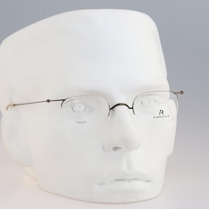 Può includere: Occhiali semi-cerchiati con montature in metallo scuro sottile e aste slanciate. Le lenti presentano la scritta Titanium a sinistra e il logo Rodenstock a destra. Questi occhiali sono esposti su una testa di manichino bianca.