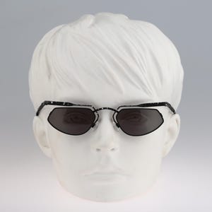 May include: Black sunglasses with a geometric frame and dark lenses. The sunglasses are on a white mannequin head.